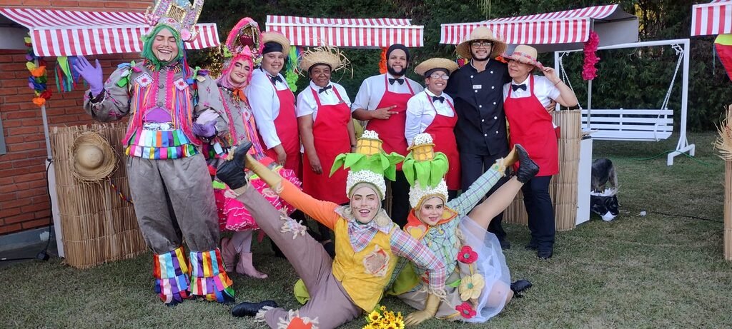 Equipe animada em frente a barraquinhas para festa junina decoradas com tecido listrado vermelho e branco, trajes típicos e comidas tradicionais